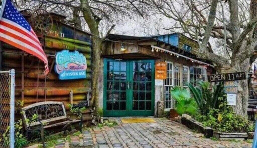 A rustic wooden building with a green door, an American flag, and a neon sign for a bar.