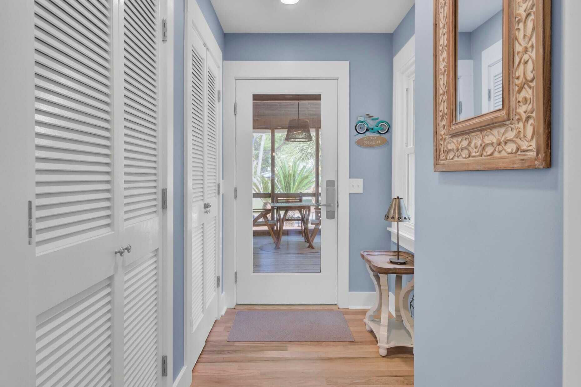 A hallway with white shutters, a wooden floor, a door leading to a room with a view, and a decorative mirror on the wall.
