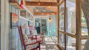 A covered porch with wooden beams, string lights, a red chair with patterned cushions, and a glass door leading to a kitchen.
