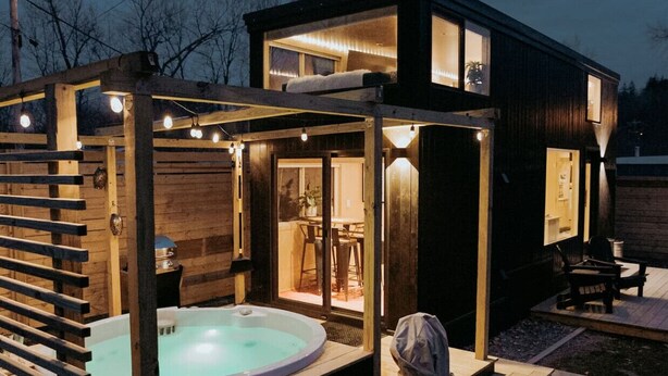 Une cabane moderne en bois, dotée d’un bain à remous, d’une terrasse en bois et d’un espace salon couvert.