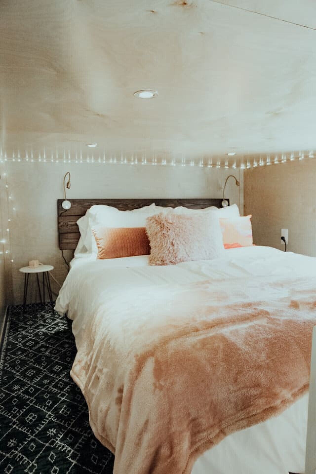 A bedroom with a wooden headboard, a bed with white and pink bedding, a small round table with a cup, and a patterned rug.