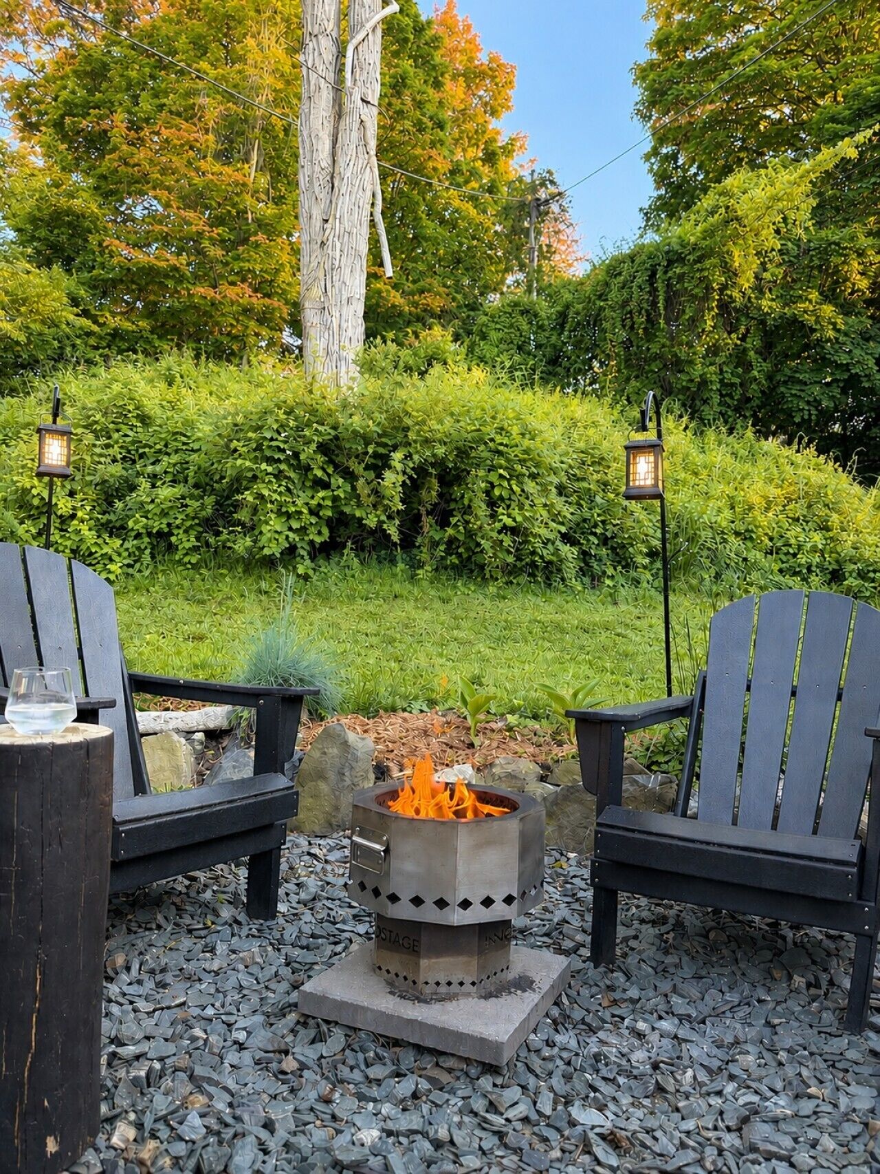 A fire pit with two Adirondack chairs and lanterns, surrounded by gravel and greenery.