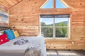 A wooden cabin bedroom with a large window, a bed with colorful pillows, and a view of a forested landscape. - Private Indoor Pool | Mtn Views | Hot Tub | Arcades | Perfect Location | Top 10% (Pigeon Forge)