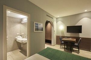 Habitación de hotel con baño, una cama, un escritorio con sillas, un televisor de pantalla plana y un cuadro en la pared.
