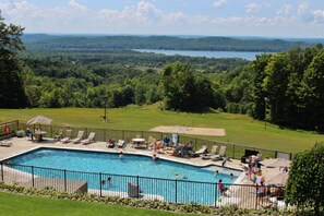 A swimming pool with lounge chairs and umbrellas, surrounded by a fence, with a view of a lake and forest in the background.