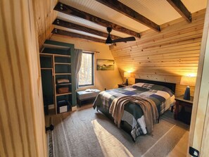 A cozy bedroom with a bed, bedside lamps, a window with curtains, a built-in shelf, and a ceiling fan.
