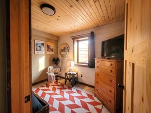 A cozy room with a wooden ceiling, a television on a dresser, a chair, a small table with a lamp, and a red and white patterned rug. - Gorgeous New A-Frame w/ Sauna & Gameroom - Minutes to Pools, Golf and Skiing (Bellaire)