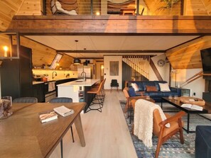 A spacious loft-style living area with a kitchen and dining area, featuring a high ceiling and wooden beams. - Gorgeous New A-Frame w/ Sauna & Gameroom - Minutes to Pools, Golf and Skiing (Bellaire)