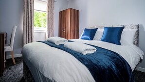 Een keurig opgemaakt bed met wit- en blauwe beddengoed, een houten garderobe en een raam met gordijnen.