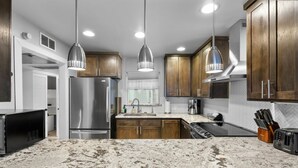 A modern kitchen with granite countertops, stainless steel appliances, wooden cabinets, pendant lighting, and a window with curtains.