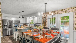 Una cocina moderna con electrodomésticos de acero inoxidable, una isla central, iluminación colgante y un comedor preparado para seis personas.