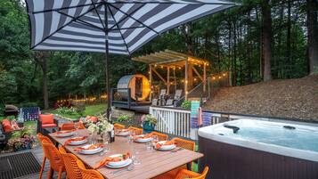 A wooden deck with a dining table set for six, surrounded by orange chairs. A hot tub is visible to the right.
