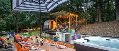 Une terrasse en bois avec une table à manger pour six personnes, entourée de chaises orange. Un jacuzzi est visible sur la droite.