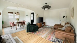 Uma sala de estar espaçosa com piso de madeira, um sofá verde, um sofá marrom, uma área de jantar com mesa e cadeiras, e uma televisão.