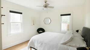 Quarto com cama, duas janelas, ventilador de teto, cadeira e luminária.
