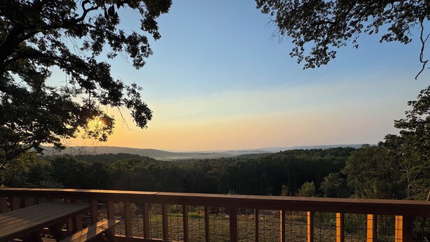 Une terrasse en bois, dotée d’une balustrade, qui offre une vue sur un paysage boisé au coucher du soleil.