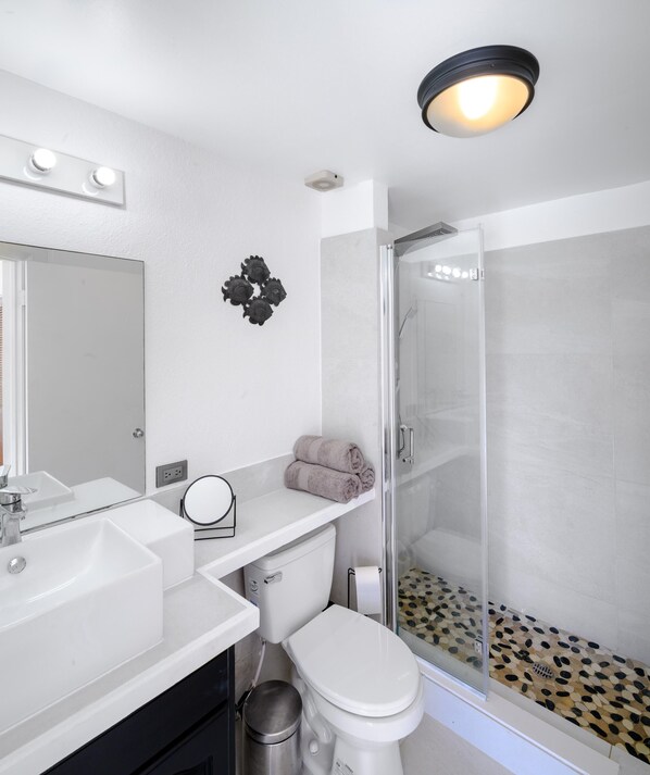 A modern bathroom with a white sink, mirror, and toilet. There is a glass shower enclosure with a pebble-patterned floor. - Wild Lotus Villa - Waterfront on Jolly Harbour (Jolly Harbour)