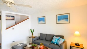 A modern living room with a grey sofa, a wooden coffee table, and a blue patterned rug. There are framed pictures on the wall, a small side table with a lamp, and a ceiling fan. - Wild Lotus Villa - Waterfront on Jolly Harbour (Jolly Harbour)
