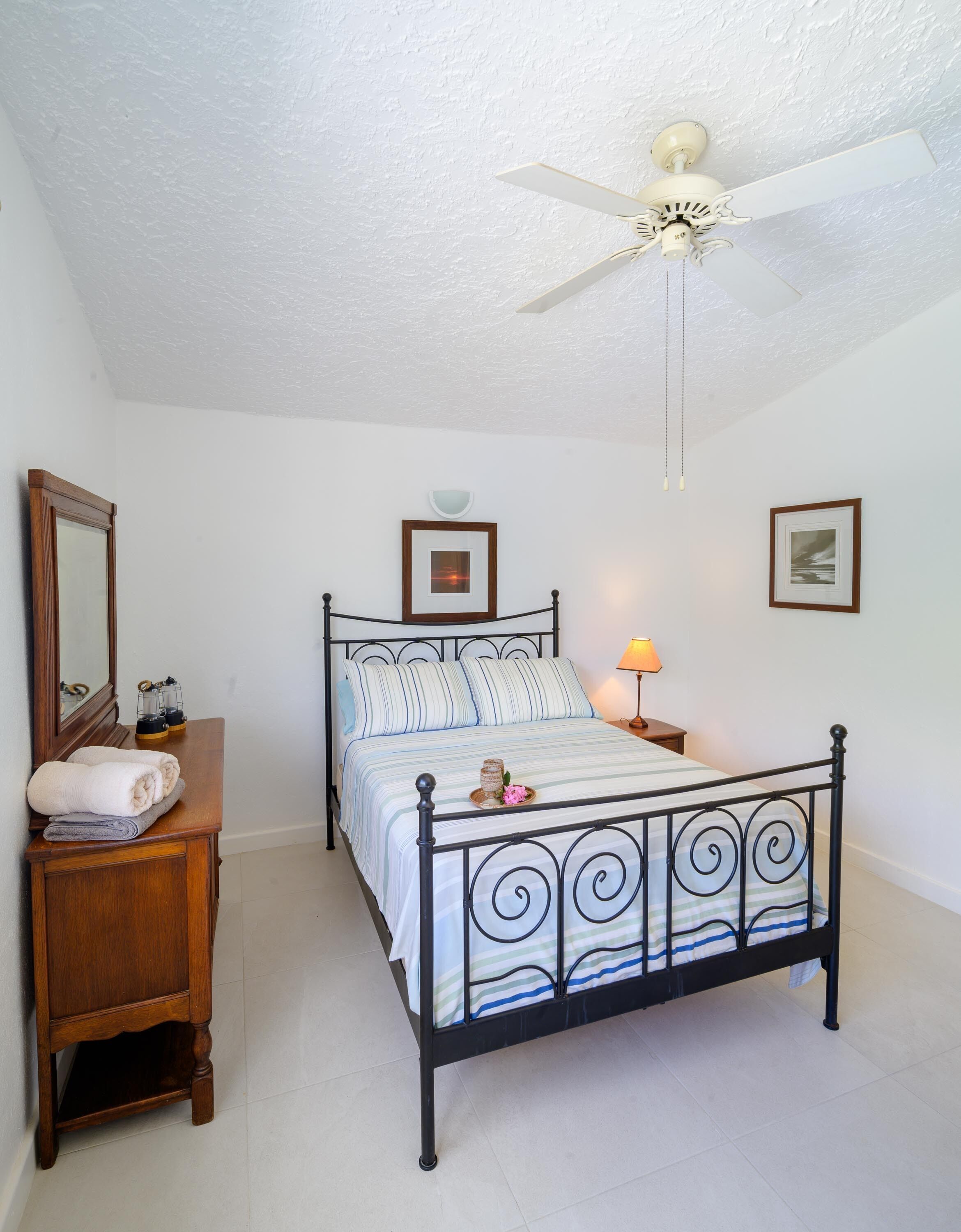 Une chambre à coucher comprenant un lit, une table de chevet, une commode, un ventilateur de plafond et des tableaux encadrés accrochés aux murs.