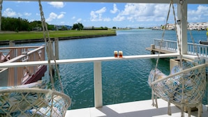 A deck with wicker chairs overlooking a body of water, a dock, and a distant shoreline. - Wild Lotus Villa - Waterfront on Jolly Harbour (Jolly Harbour)