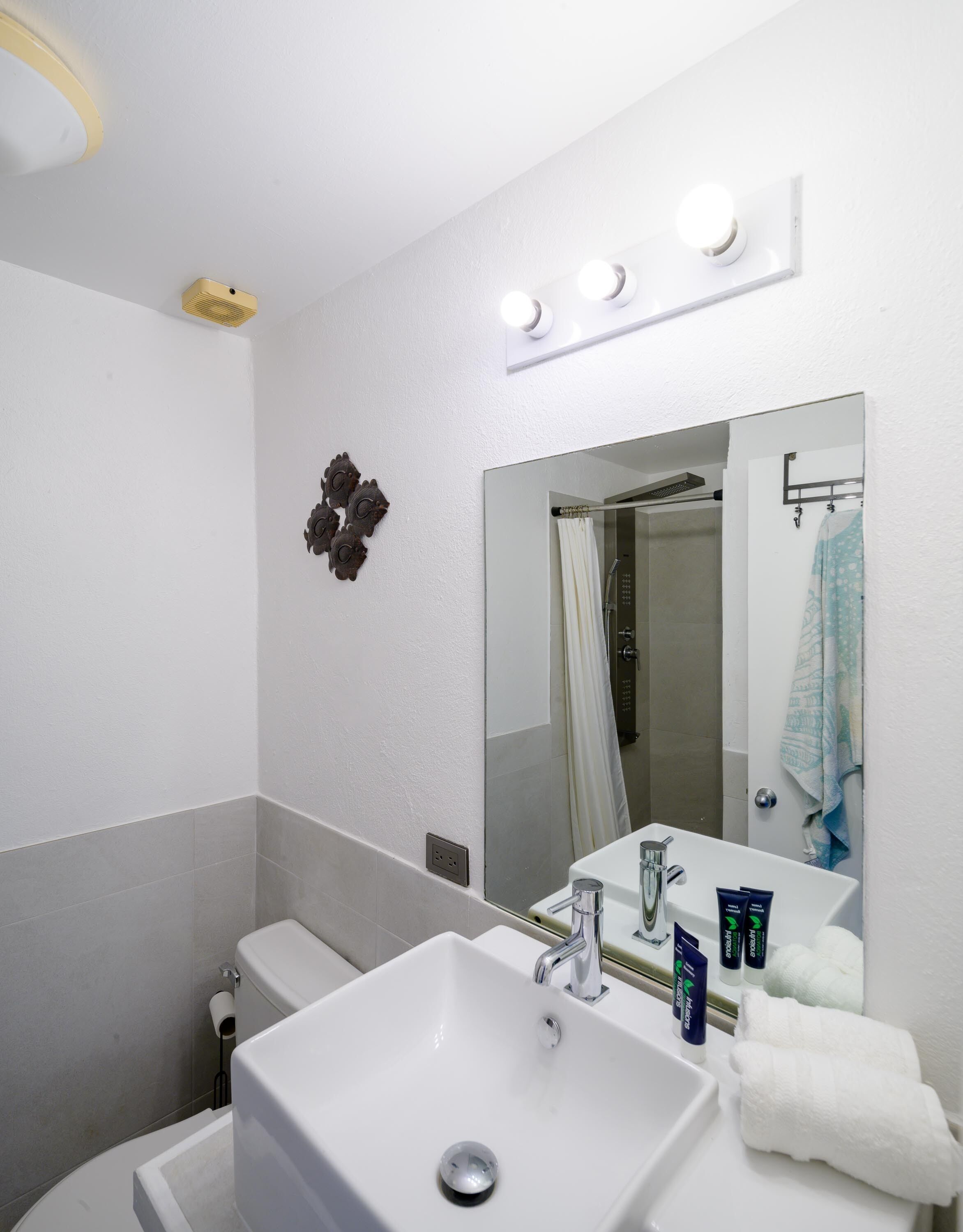 A bathroom with a white sink, mirror, and shower. There are three lights above the mirror and a ceiling fan.