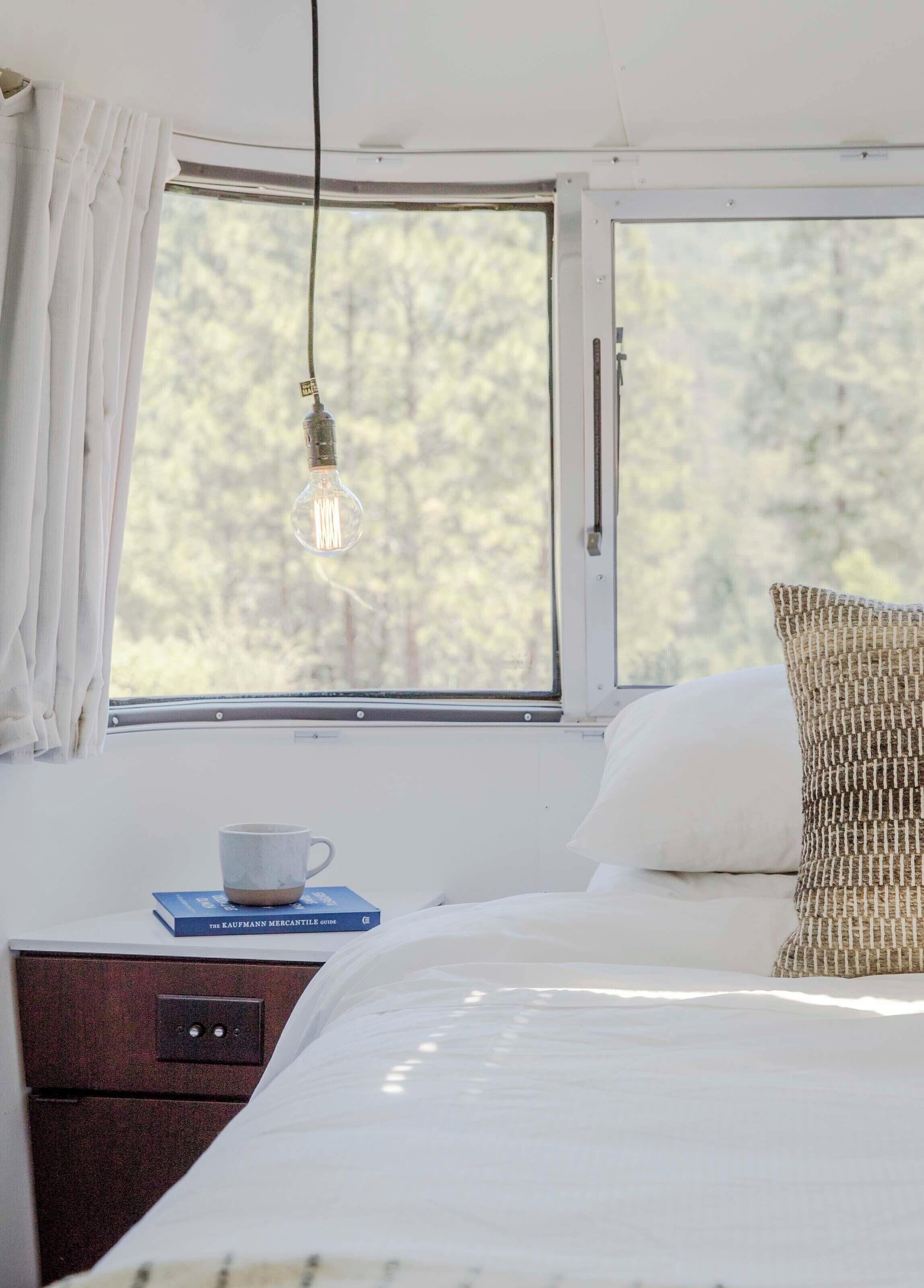 Un dormitorio con cama, ventana con cortinas, bombilla colgante, mesita de noche con taza y libro, y almohada texturizada.