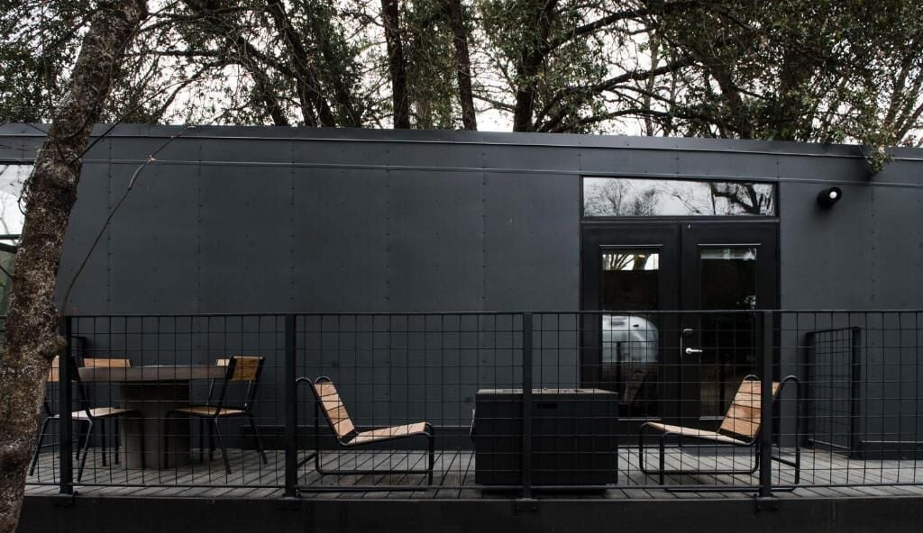 Un edificio moderno con fachada oscura, un patio amurallado con mobiliario y una puerta visible.
