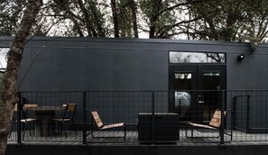 A modern building with a dark exterior, a fenced patio area with seating, and a visible door.
