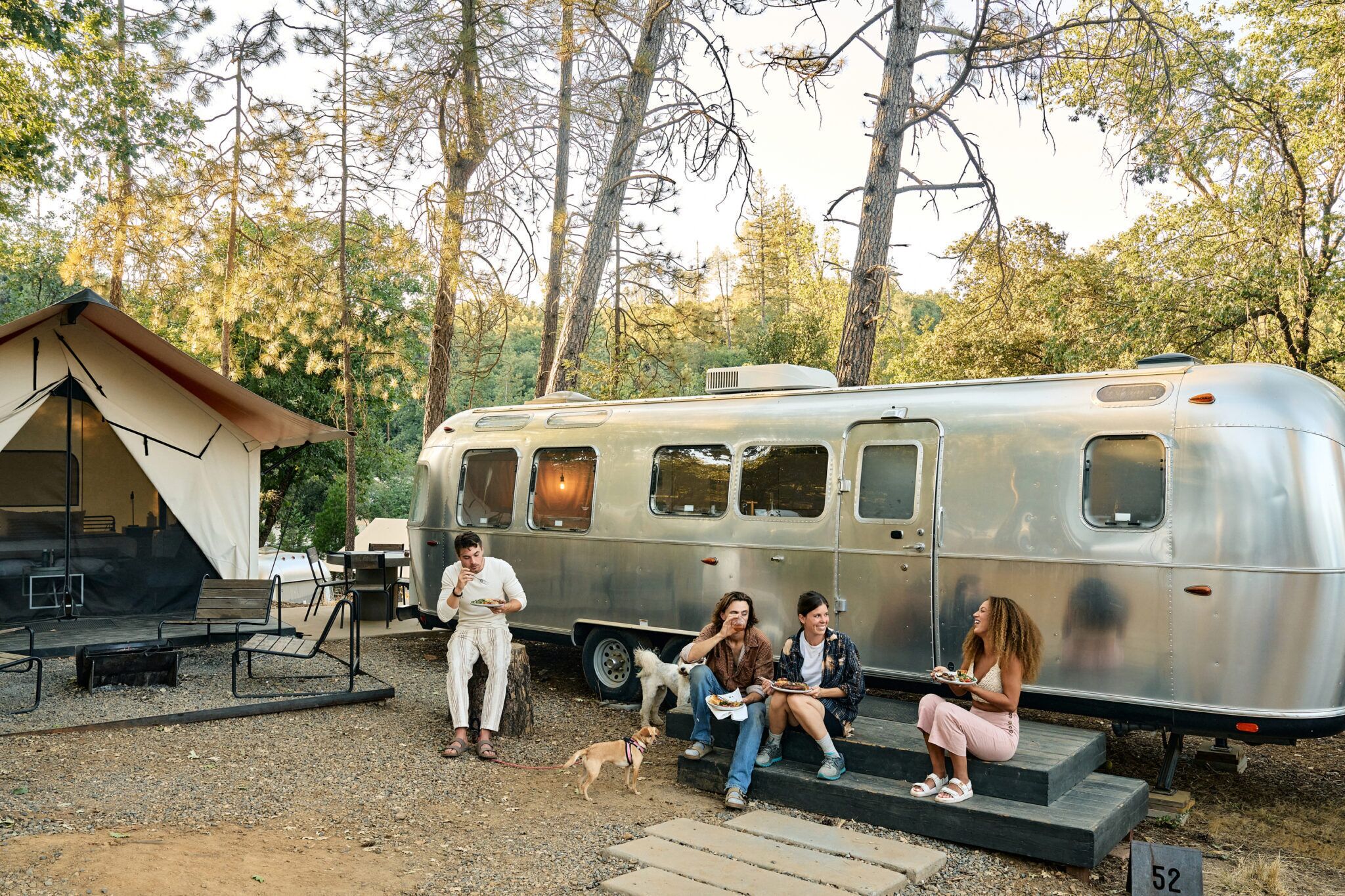 A group of people sitting on steps next to a vintage trailer, with a tent and outdoor seating area in the background.