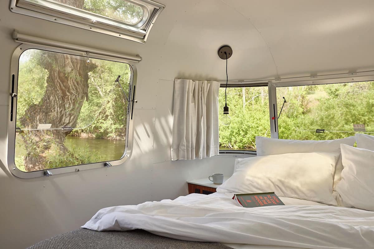 Una cabaña con una cama, un ventanal con vistas a un bosque y una mesita con un libro y una taza.