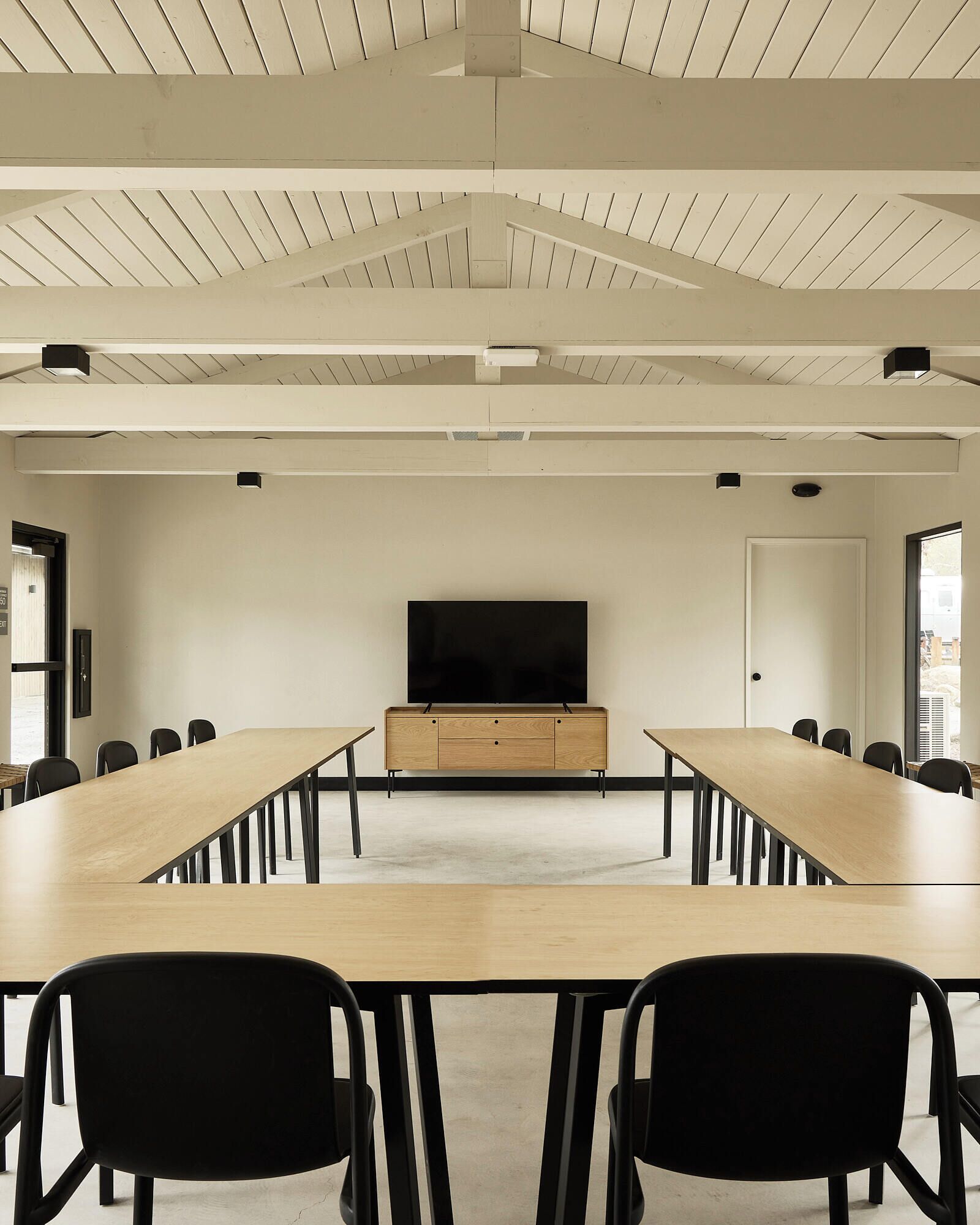 A conference room with a long table, black chairs, a television on a wooden cabinet, and a white ceiling with wooden beams.