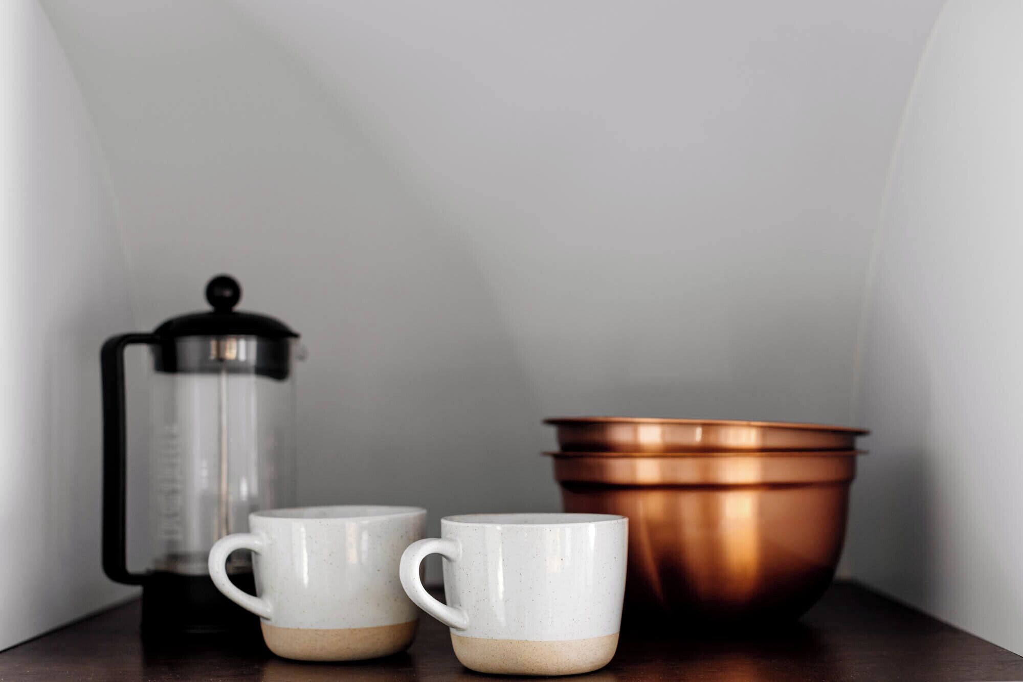 Dos tazas blancas con diseños moteados, una prensa francesa negra y plateada, y un recipiente de color cobre sobre una superficie de madera.