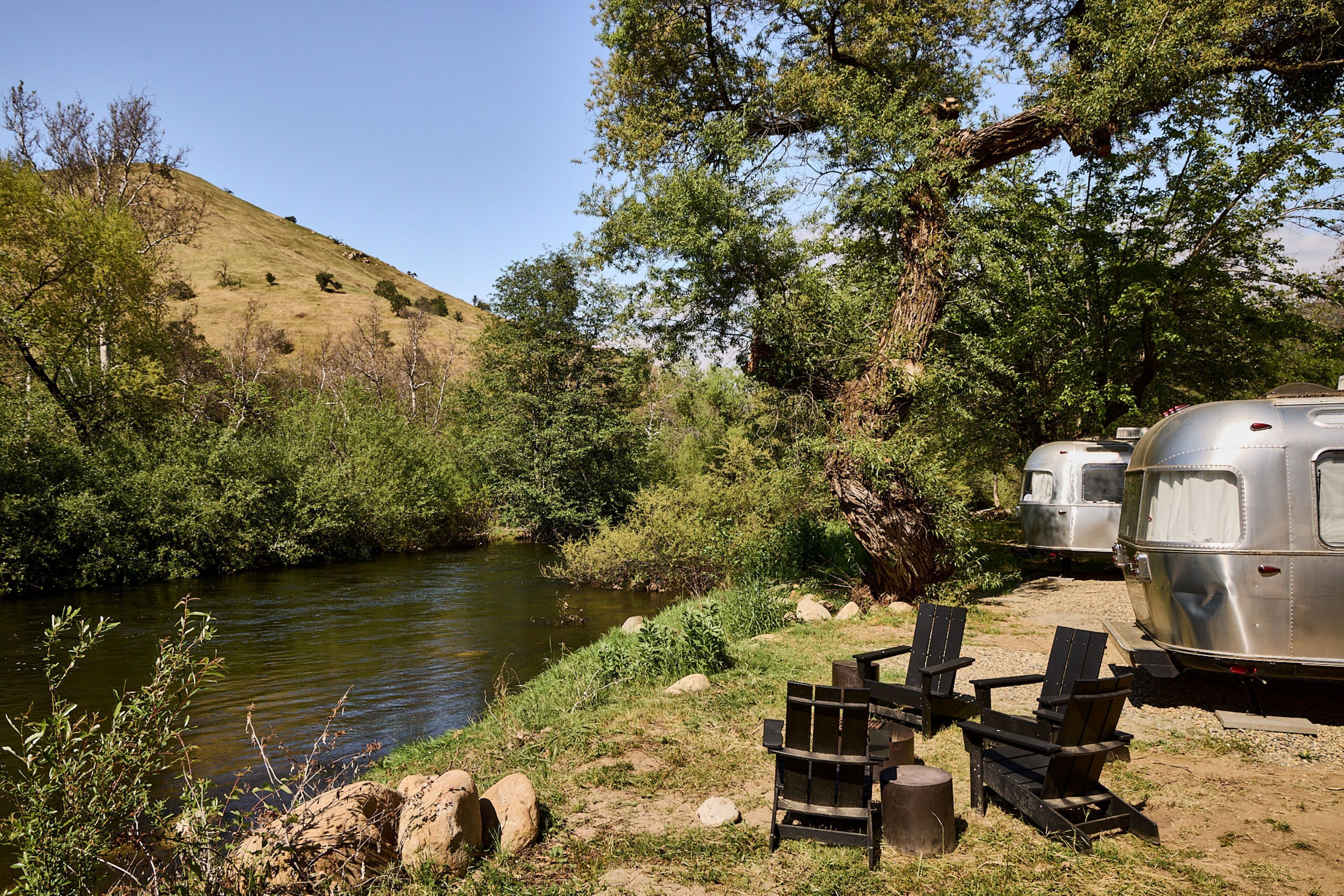 A campsite with two trailers by a river, surrounded by trees and hills.