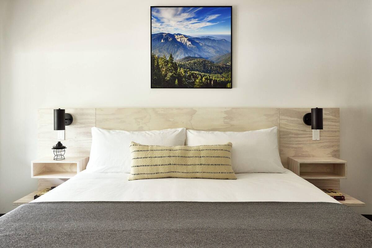 Un dormitorio con cabecera de madera, ropa de cama blanca, un cuadro de un paisaje montañoso y dos mesitas de noche con lámparas.