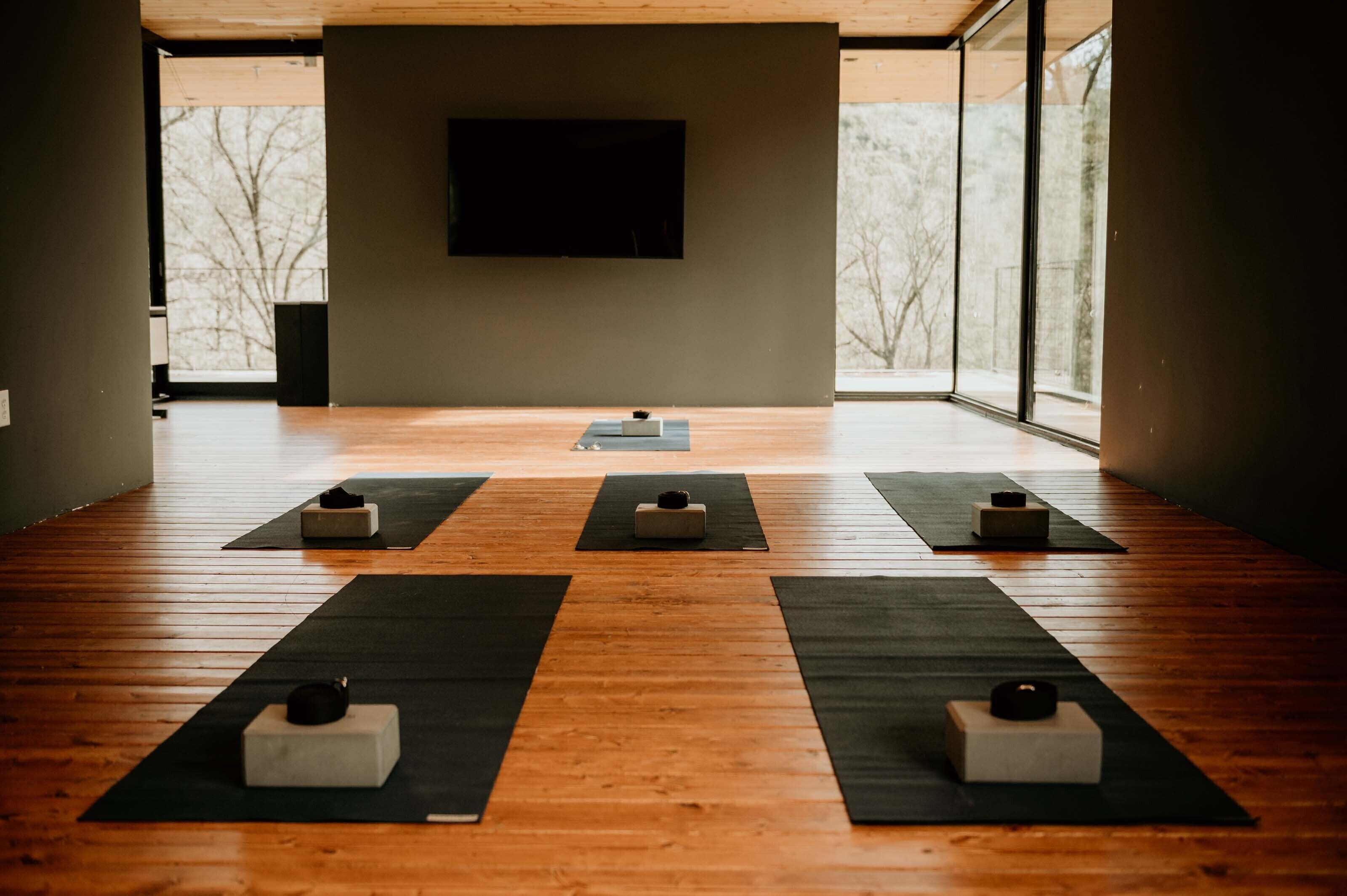 Un estudio de yoga con suelo de madera, tapetes negros y bloques dispuestos para la práctica.