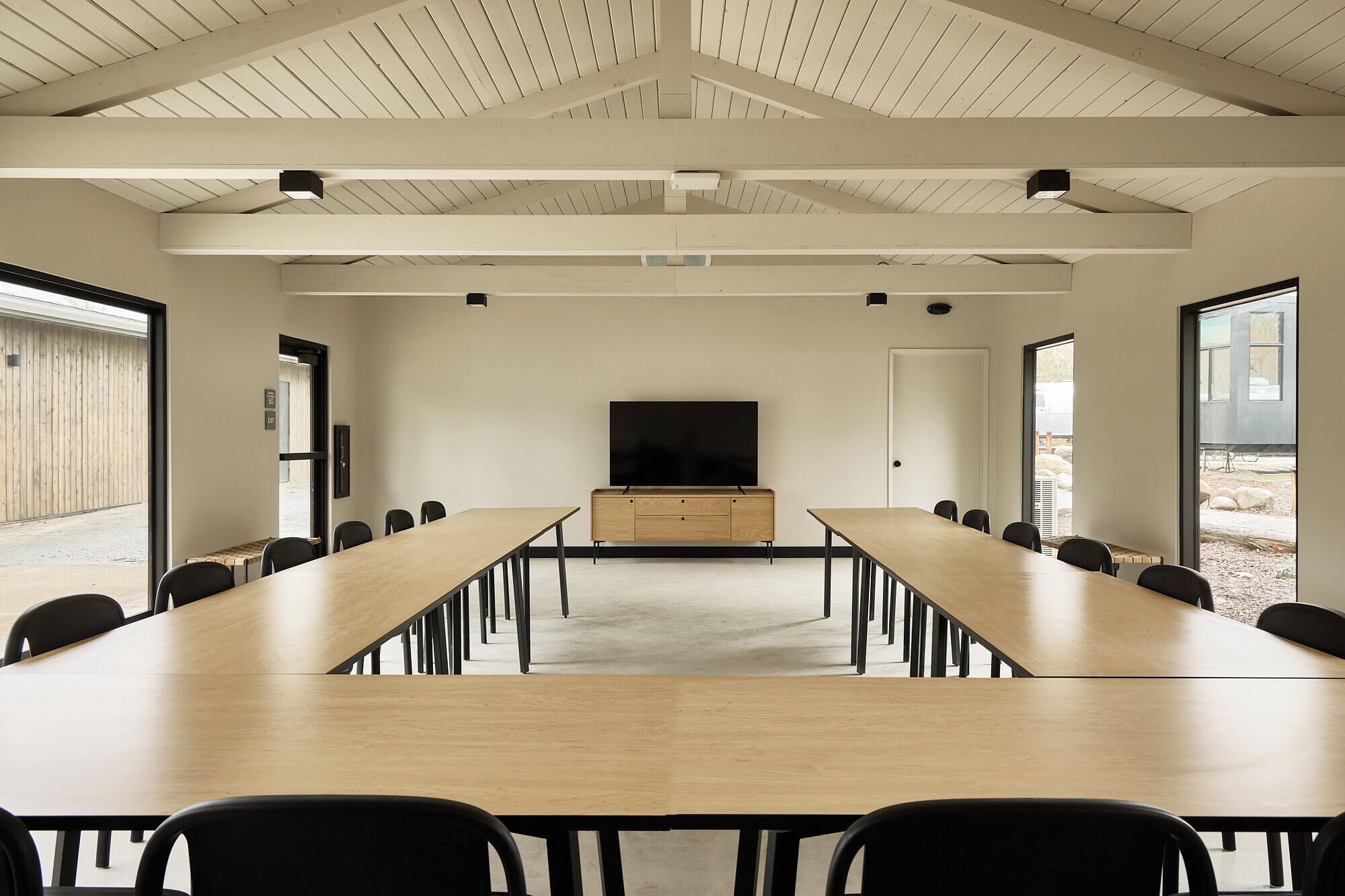 A conference room with long tables and chairs, a large flat-screen TV, and a high ceiling with exposed beams.