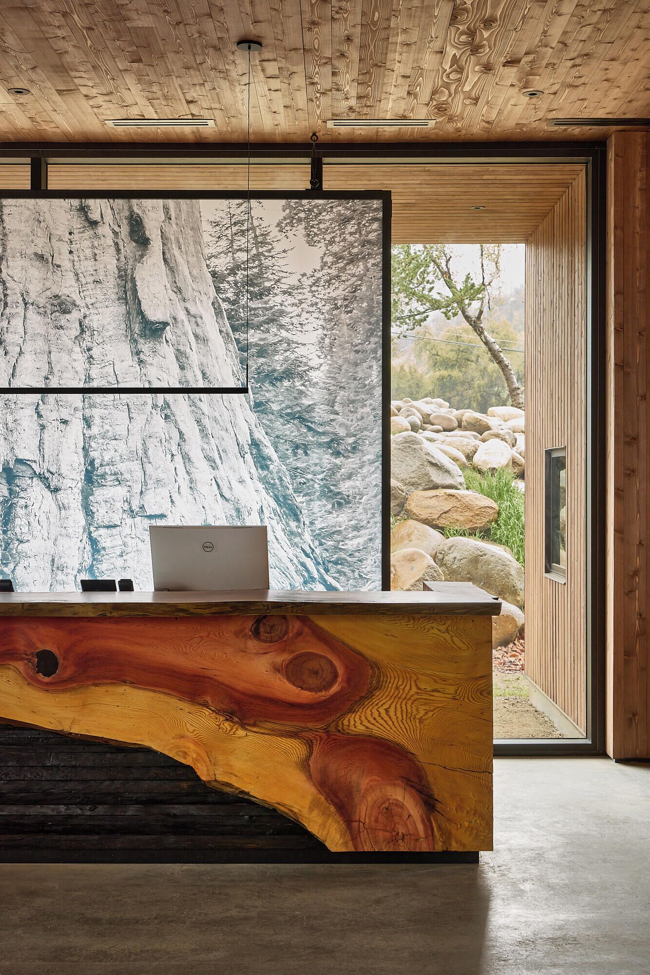 A reception desk with a wooden design, a laptop, and a large window with a view of rocks and trees.
