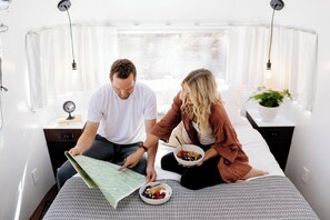 A couple sitting on a bed in a well-lit room with a map, bowls of cereal, and a plant.