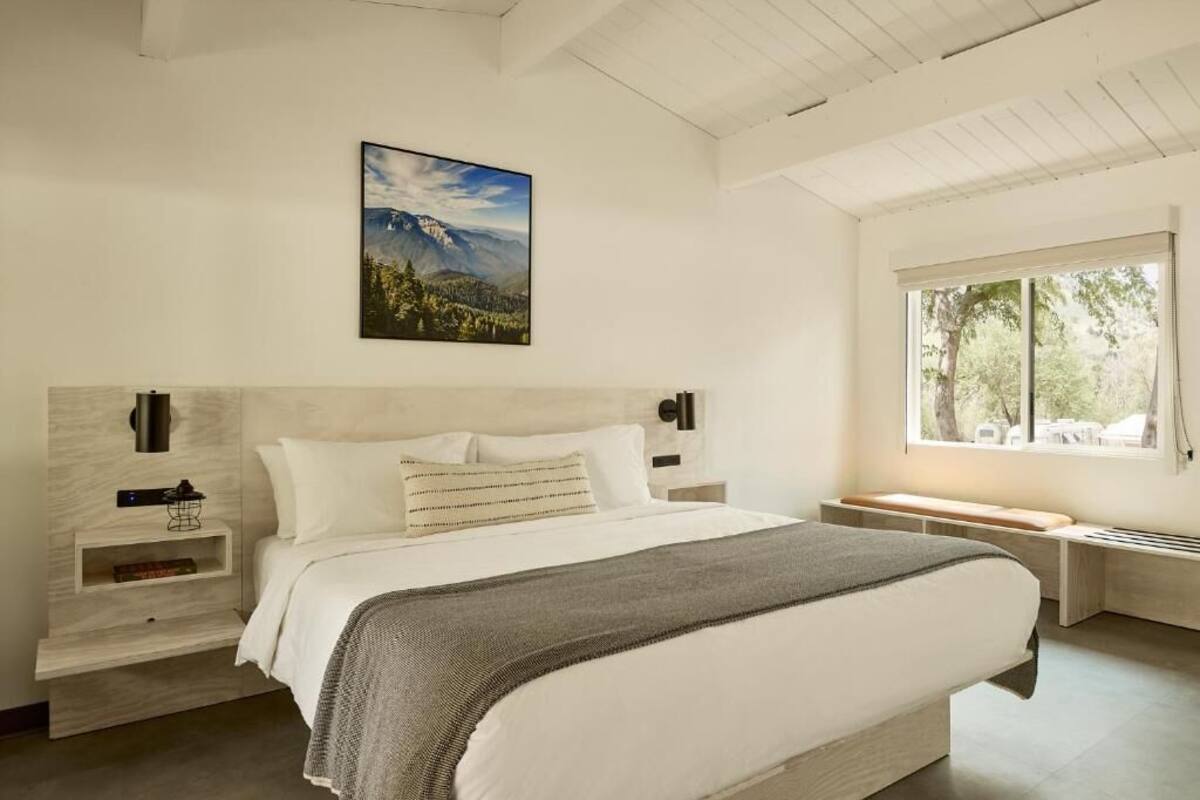 Un dormitorio con cama, mesitas de noche, una ventana y un cuadro en la pared.