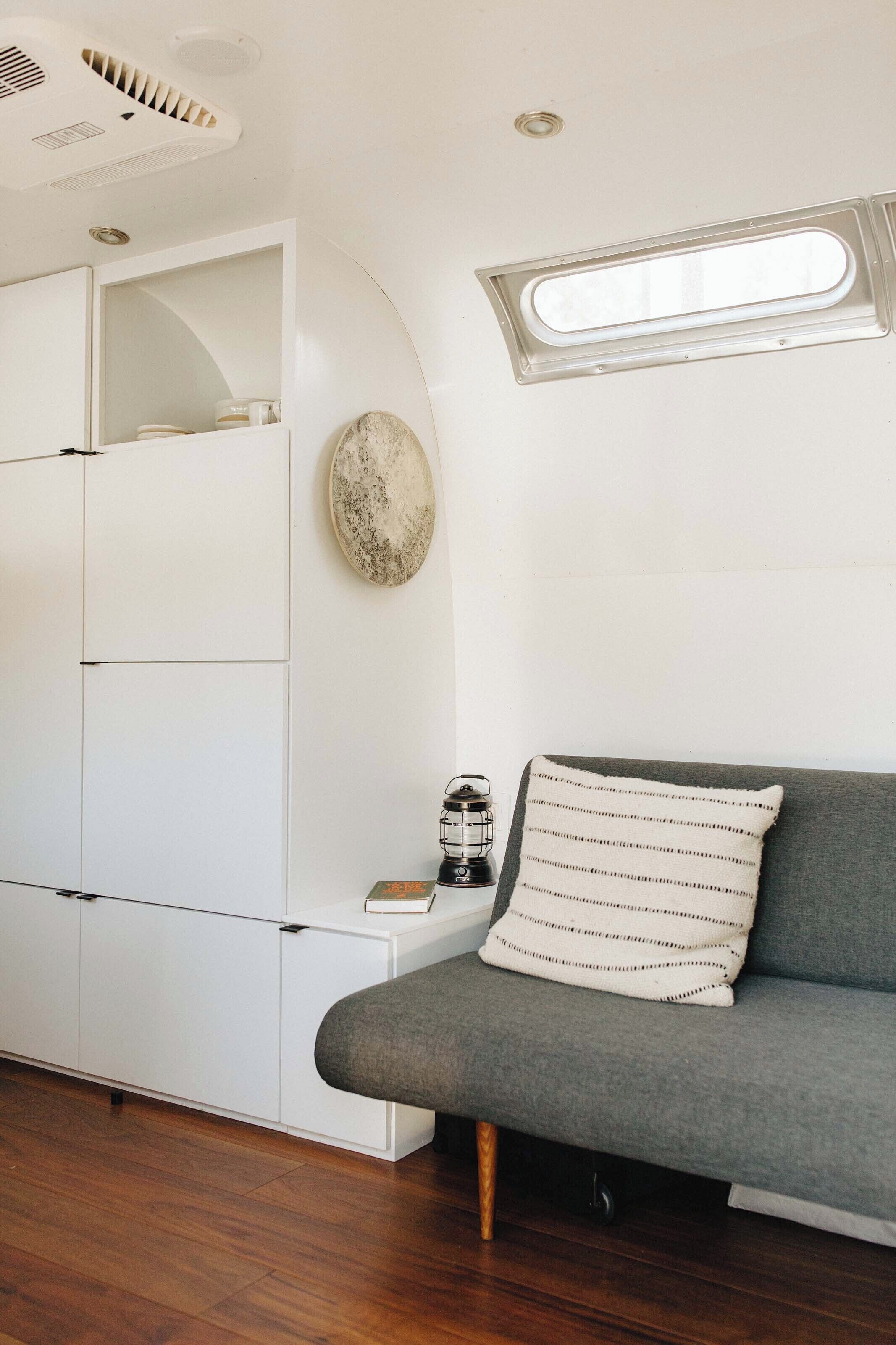 A compact living space with a grey sofa, white cabinetry, a small table with a lantern, and a window with a view.