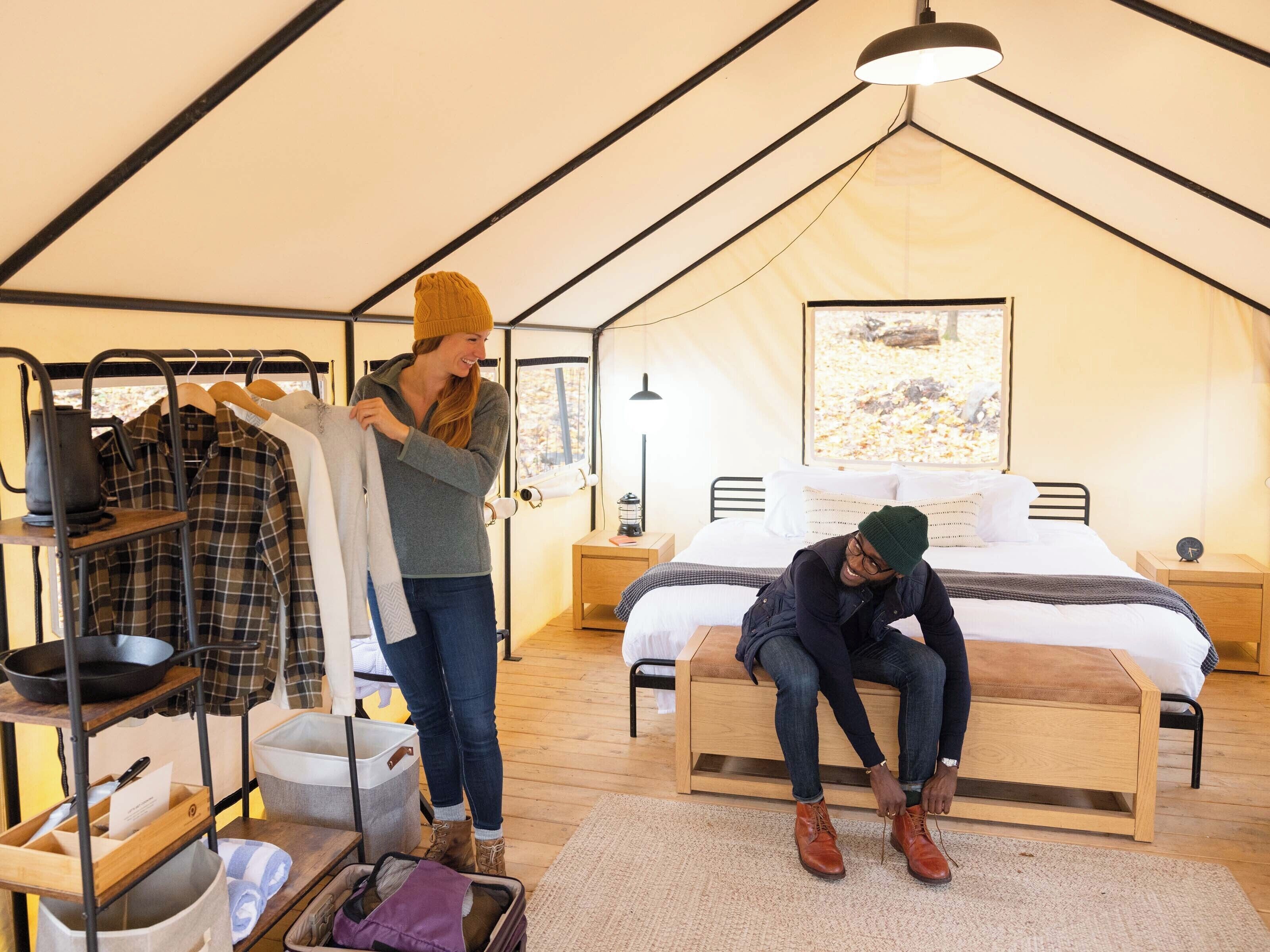 Una tienda de campaña con una cama, un banco de madera, un estante con ropa y una maleta.