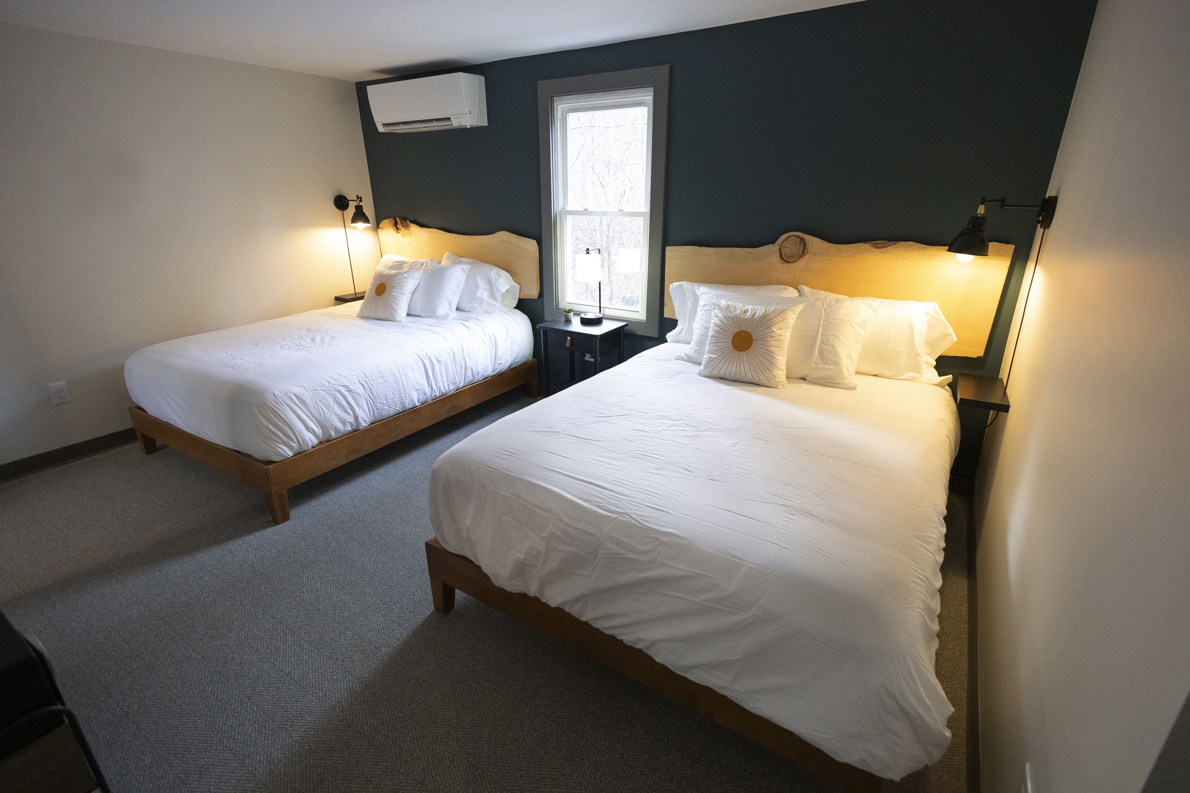A hotel room with two single beds, a nightstand with a lamp, a window with natural light, and an air conditioning unit on the wall.