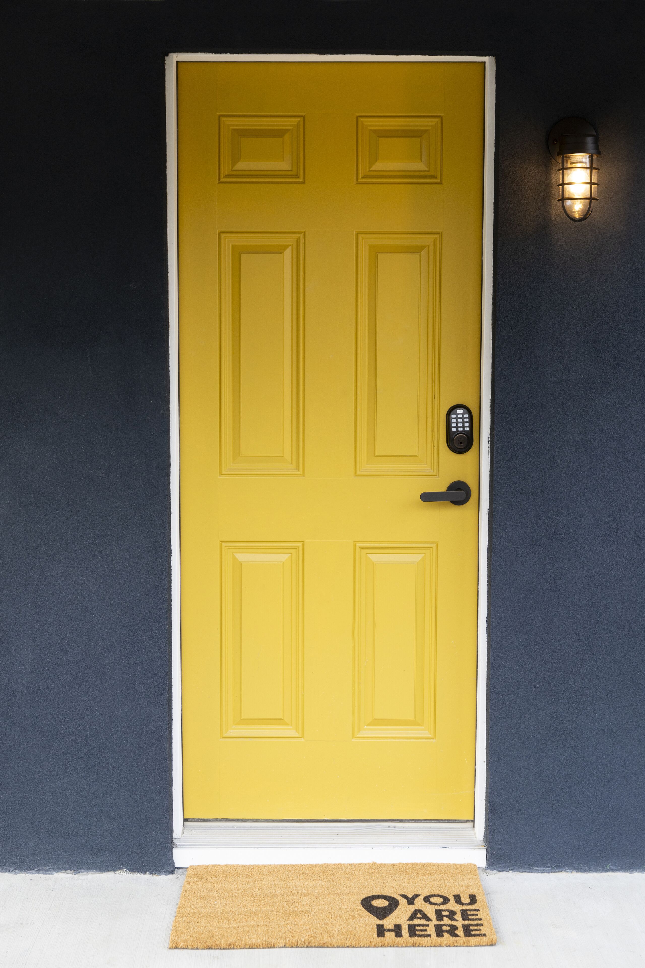 A yellow door with a keypad lock, a doormat with "YOU ARE HERE" text, and a wall-mounted light fixture.