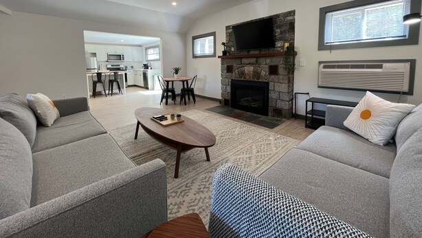 A modern living room with grey sofas, a wooden coffee table, a stone fireplace, and a kitchen in the background. - East Rock Inn (Great Barrington)