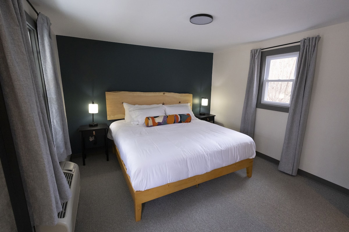 Un dormitorio ordenado con una cama de madera, una mesita de noche, una lámpara y una ventana con cortinas.