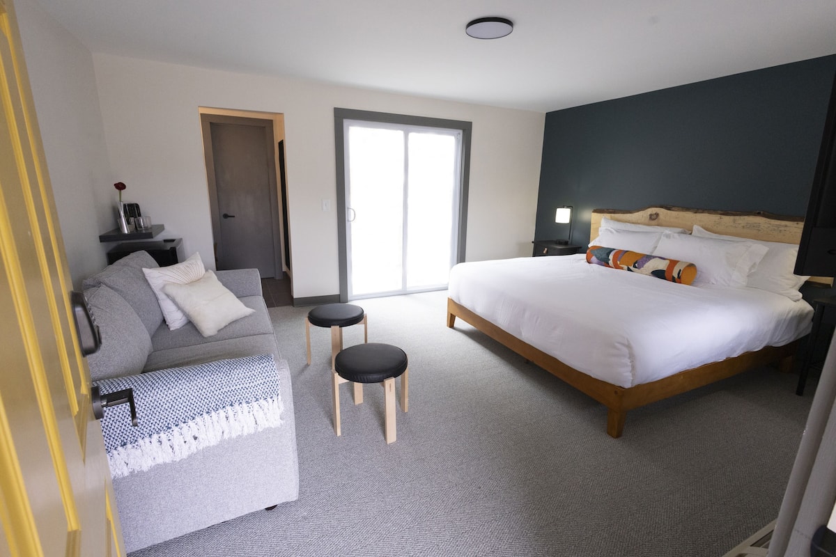 Un dormitorio con cama, sofá, dos taburetes, una mesita de noche, lámpara, puerta y ventana.