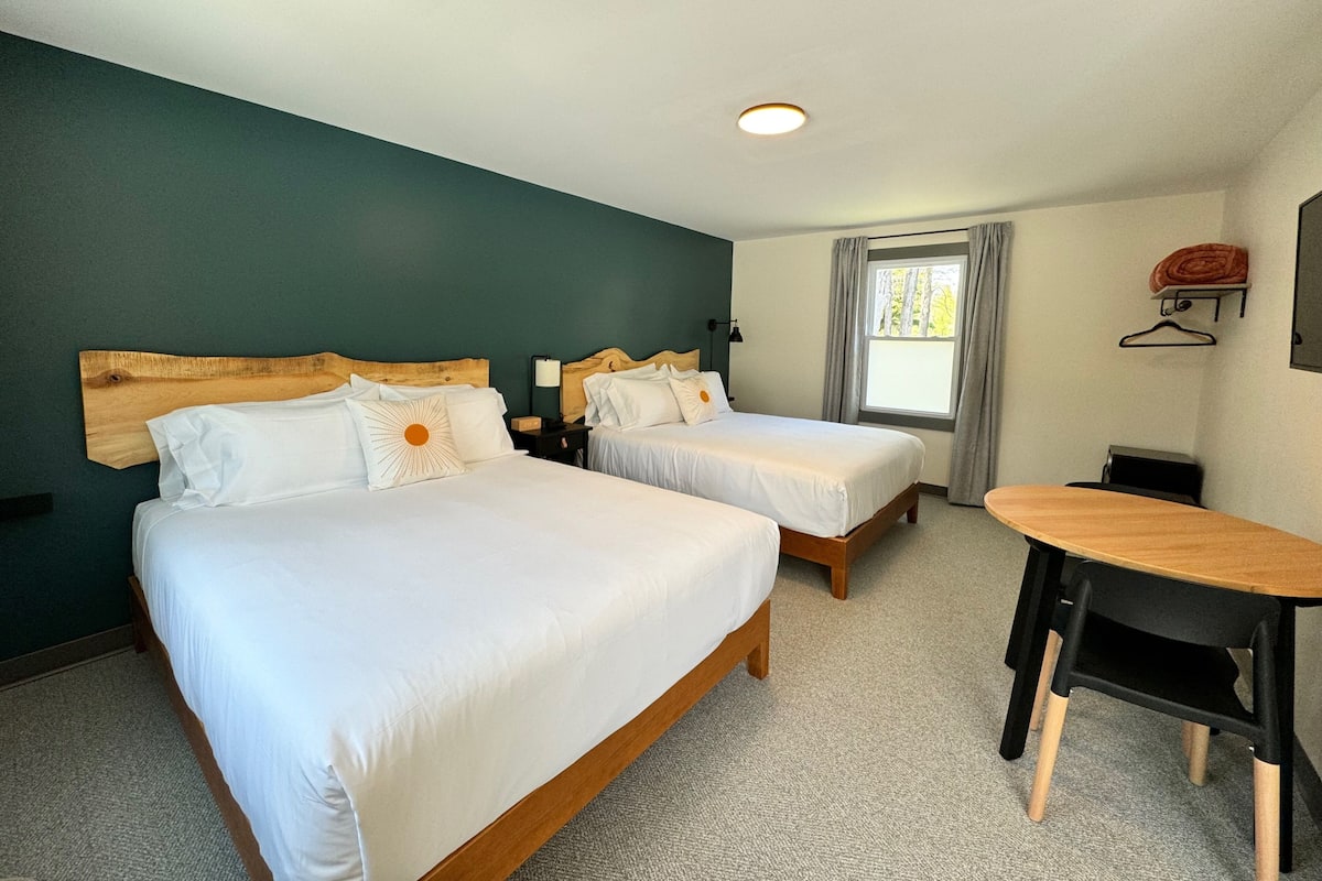 Habitación de hotel con dos camas, un escritorio, una silla, una ventana con cortinas y un perchero en la pared.