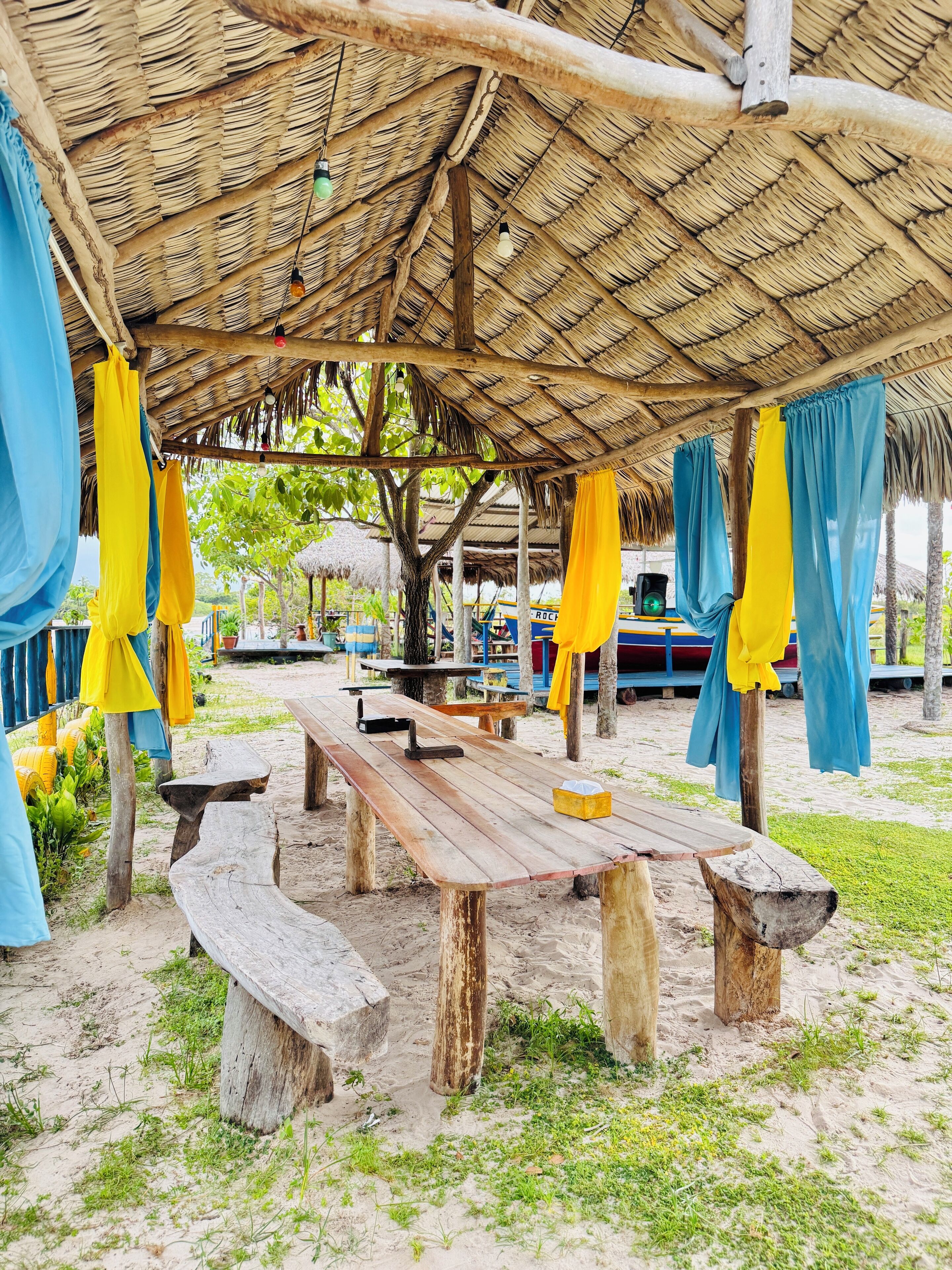 A beachside pavilion with wooden benches and a thatched roof, surrounded by greenery.