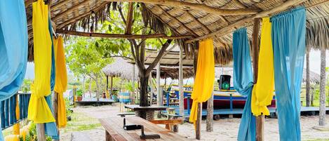 A beachside pavilion with wooden benches and a thatched roof, surrounded by greenery.