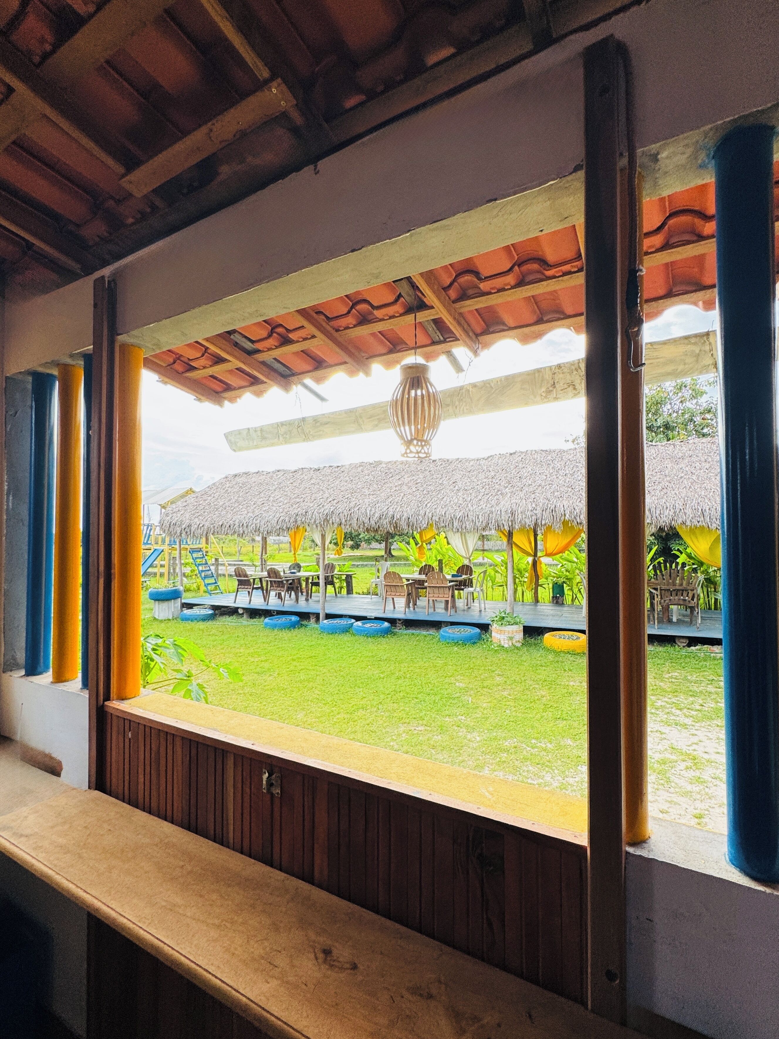 Vista desde una ventana que muestra una habitación con banca de madera, un espacio al aire libre con techo de paja, sillas y mesas, y una lámpara colgante.